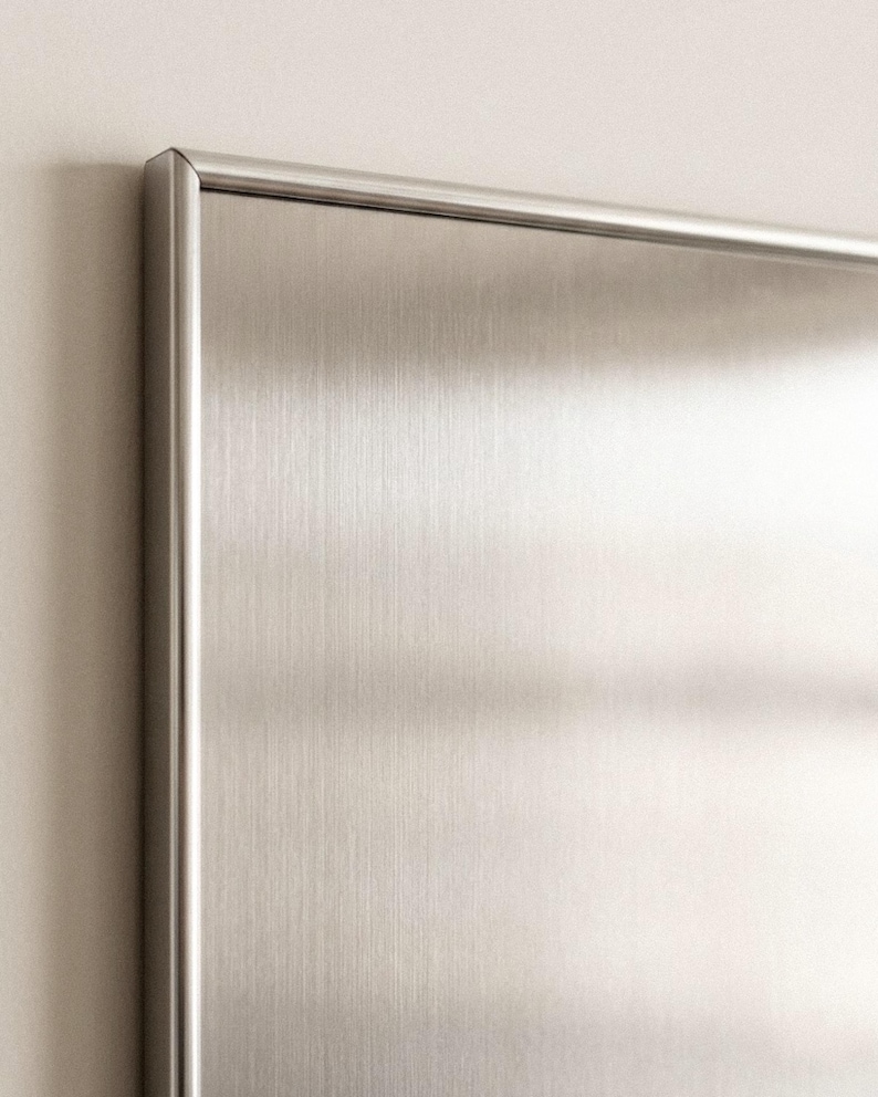Puede incluir: Primer plano de un panel de acero inoxidable cepillado con un marco met&aacute;lico delgado. La superficie tiene un patr&oacute;n de vetas verticales, que refleja la luz. El marco tiene un borde redondeado. El panel est&aacute; montado en una pared de color claro.