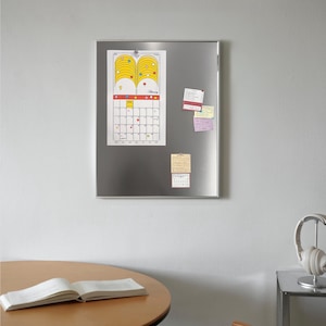 Puede incluir: Un tablero magn&eacute;tico de metal plateado con un calendario y notas adhesivas adjuntas. El calendario tiene un dise&ntilde;o amarillo y naranja en la parte superior y una cuadr&iacute;cula para los d&iacute;as del mes. Una mesa redonda de madera con un libro abierto est&aacute; en primer plano.