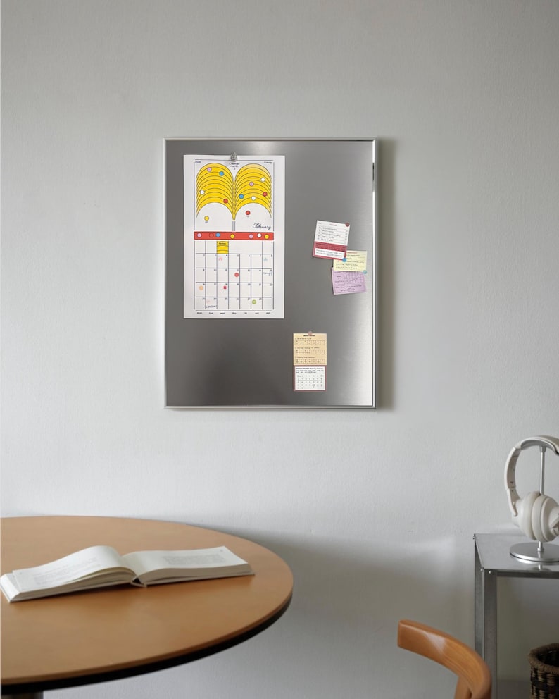 Puede incluir: Un tablero magn&eacute;tico de metal plateado con un calendario y notas adhesivas adjuntas. El calendario tiene un dise&ntilde;o amarillo y naranja en la parte superior y una cuadr&iacute;cula para los d&iacute;as del mes. Una mesa redonda de madera con un libro abierto est&aacute; en primer plano.