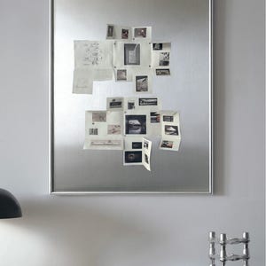 Puede incluir: Un gran tablero magn&eacute;tico rectangular plateado con varios fotos y bocetos peque&ntilde;os adjuntos. El tablero est&aacute; montado en una pared gris claro. Una taza y un platillo son visibles en una mesa en primer plano.
