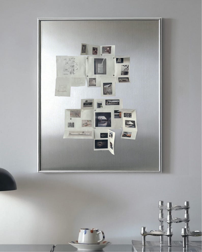 Puede incluir: Un gran tablero magn&eacute;tico rectangular plateado con varios fotos y bocetos peque&ntilde;os adjuntos. El tablero est&aacute; montado en una pared gris claro. Una taza y un platillo son visibles en una mesa en primer plano.