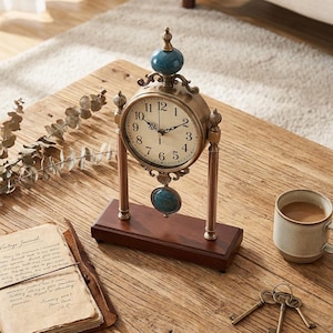 May include: An antique-style clock with a bronze-colored frame and a cream-colored clock face. The clock has a dark teal-colored sphere on top and a matching oval-shaped pendant. The clock sits on a dark brown wooden base.