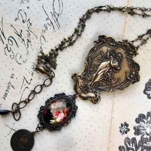 May include: A bronze necklace with a large pendant featuring a woman in a flowing dress. The pendant is surrounded by intricate floral designs. The necklace also has a smaller pendant with a floral design and a black background.