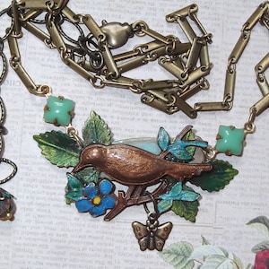 May include: A bronze bird perched on a branch with green leaves and blue flowers. The bird is part of a necklace with a green stone pendant and a gold chain.