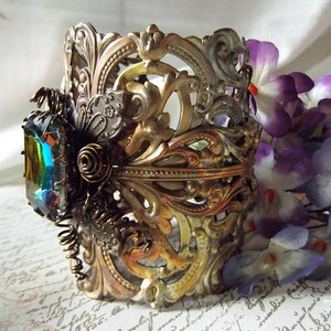 May include: A gold-toned metal cuff bracelet with an intricate, ornate design. The bracelet features a large, iridescent gemstone set in the center. The bracelet is accented with a decorative, coiled wire.