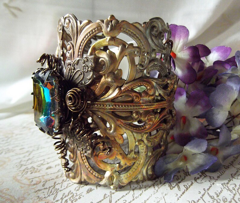 May include: A gold-toned metal cuff bracelet with an intricate, ornate design. The bracelet features a large, iridescent gemstone set in the center. The bracelet is accented with a decorative, coiled wire.