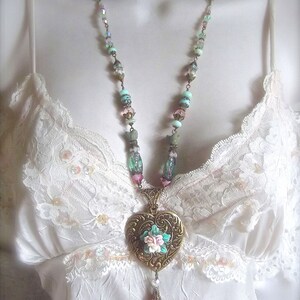 Puede incluir: Un collar de estilo vintage con un colgante en forma de coraz&oacute;n. El colgante de color bronce presenta un dise&ntilde;o floral con acentos rosas, blancos y turquesas. El collar tiene una cadena de cuentas con cuentas de color pastel y un dije colgante.