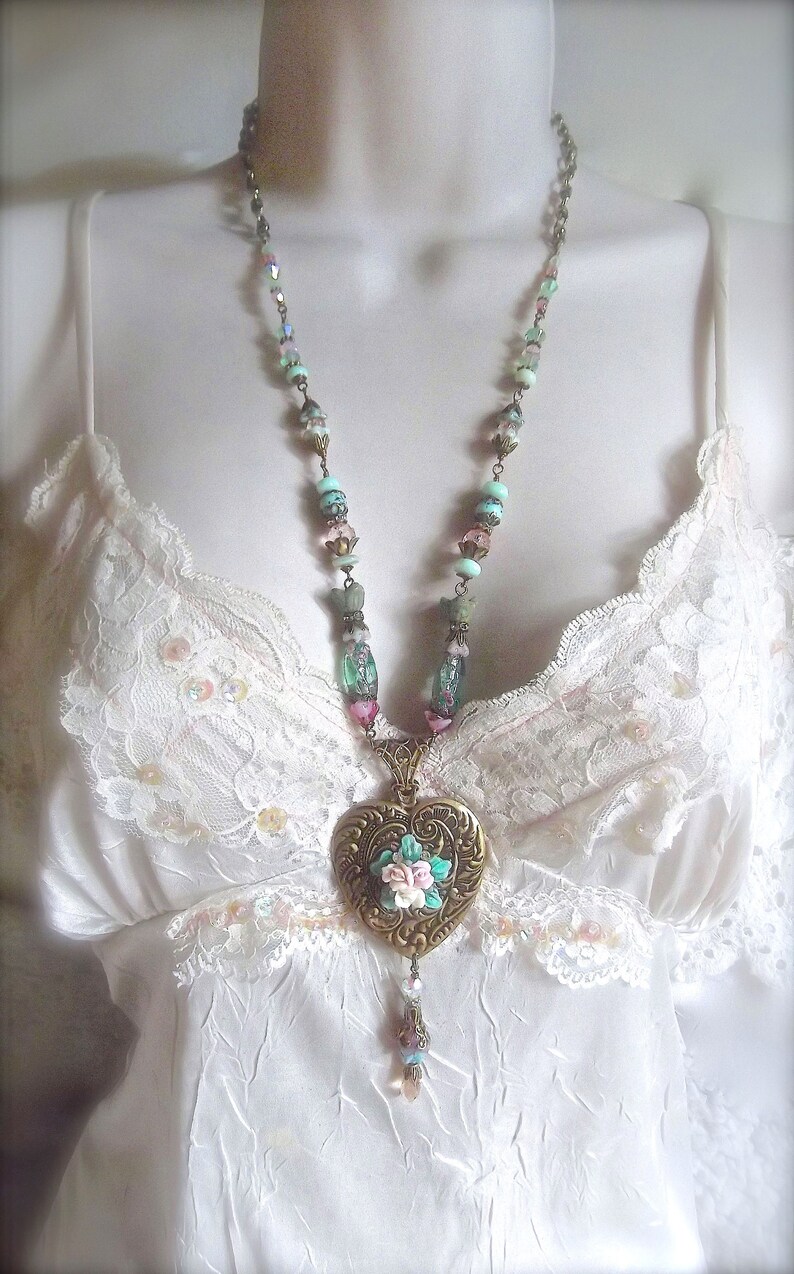 Puede incluir: Un collar de estilo vintage con un colgante en forma de coraz&oacute;n. El colgante de color bronce presenta un dise&ntilde;o floral con acentos rosas, blancos y turquesas. El collar tiene una cadena de cuentas con cuentas de color pastel y un dije colgante.