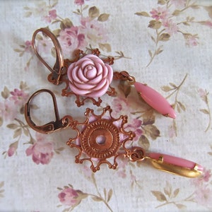 May include: A pair of copper earrings with pink rose-shaped accents. The earrings feature intricate filigree designs and dangle from a hook.