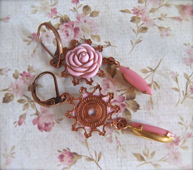May include: A pair of copper earrings with pink rose-shaped accents. The earrings feature intricate filigree designs and dangle from a hook.
