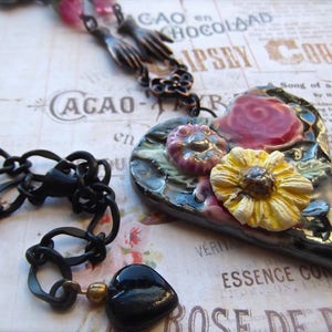 Pode incluir: Um colar de corrente preta com um pingente em forma de cora&ccedil;&atilde;o. O pingente &eacute; decorado com flores e tem um esmalte preto, verde e dourado. As flores s&atilde;o rosa, amarelas e brancas.