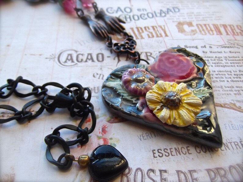 Pode incluir: Um colar de corrente preta com um pingente em forma de cora&ccedil;&atilde;o. O pingente &eacute; decorado com flores e tem um esmalte preto, verde e dourado. As flores s&atilde;o rosa, amarelas e brancas.