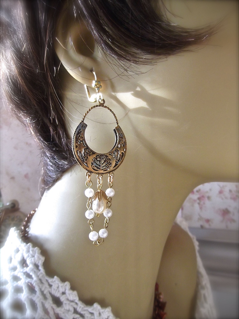 Puede incluir: Pendientes de tono dorado con un gran dise&ntilde;o circular adornado y una perla colgante. Los pendientes est&aacute;n en un maniqu&iacute; blanco.