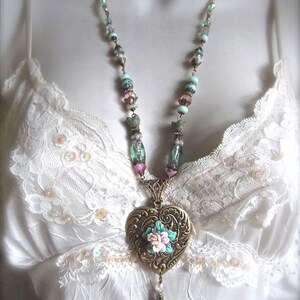 Puede incluir: Un collar de estilo vintage con un colgante en forma de coraz&oacute;n. El colgante es de color bronce con un dise&ntilde;o floral en el centro. La cadena tiene cuentas de colores pastel y un dije colgante. El collar se muestra sobre un fondo blanco.