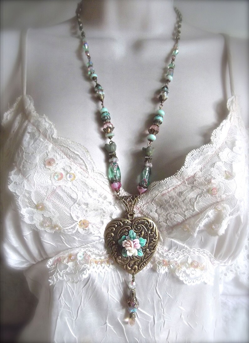 Puede incluir: Un collar de estilo vintage con un colgante en forma de coraz&oacute;n. El colgante es de color bronce con un dise&ntilde;o floral en el centro. La cadena tiene cuentas de colores pastel y un dije colgante. El collar se muestra sobre un fondo blanco.