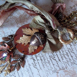 May include: A brown, gold and green necklace with a large acorn pendant and a chain made of metal links. The necklace is adorned with fabric scraps in shades of brown, green and pink.