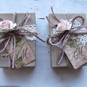 Puede incluir: Dos peque&ntilde;as cajas de regalo marrones atadas con cuerda y decoradas con encaje, flores rosas y una etiqueta que dice "M".