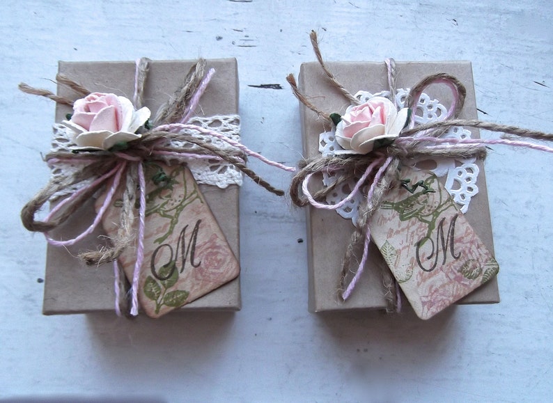 Puede incluir: Dos peque&ntilde;as cajas de regalo marrones atadas con cuerda y decoradas con encaje, flores rosas y una etiqueta que dice "M".