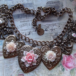 May include: A bronze chain necklace with multiple heart-shaped charms. Each charm features a raised, ornate design and a delicate pink flower. The chain is made of linked rings.