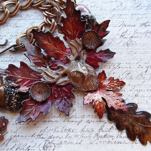 May include: A necklace with a bronze deer head pendant surrounded by fall-colored leaves. The leaves are made of metal and have a shiny, metallic finish. The necklace is attached to a chain with a lobster clasp.