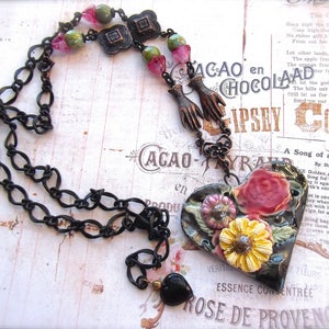 Pode incluir: Um colar de corrente preta com um pingente em forma de cora&ccedil;&atilde;o. O pingente &eacute; feito de argila preta e apresenta um design floral com flores rosa, amarelas e verdes. O colar tamb&eacute;m tem uma conta preta e v&aacute;rias contas de vidro verdes e rosas.
