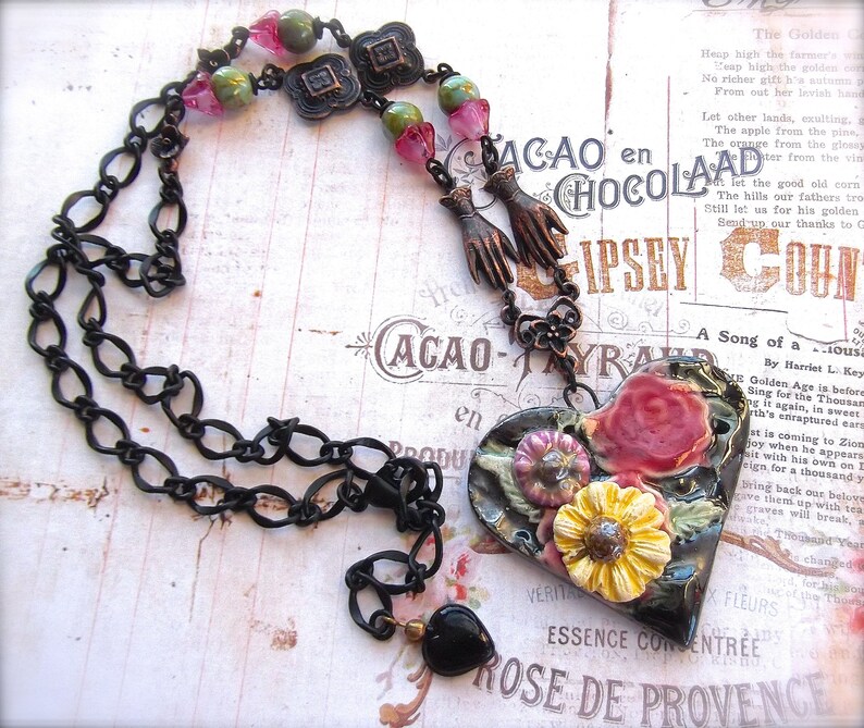 Pode incluir: Um colar de corrente preta com um pingente em forma de cora&ccedil;&atilde;o. O pingente &eacute; feito de argila preta e apresenta um design floral com flores rosa, amarelas e verdes. O colar tamb&eacute;m tem uma conta preta e v&aacute;rias contas de vidro verdes e rosas.