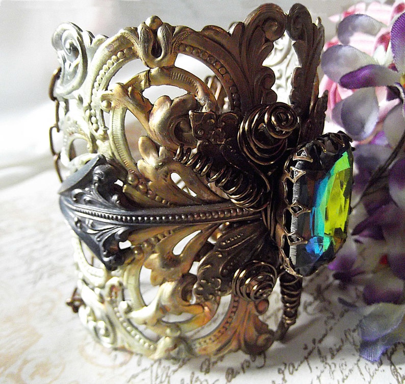 May include: A gold and silver ornate bracelet with a large iridescent green gemstone. The bracelet is made of metal and has a delicate, intricate design.