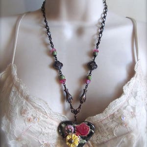Pode incluir: Um colar de corrente preta com contas verdes e rosa e um pingente em forma de cora&ccedil;&atilde;o. O pingente &eacute; decorado com um desenho floral em tons de rosa, amarelo e verde.