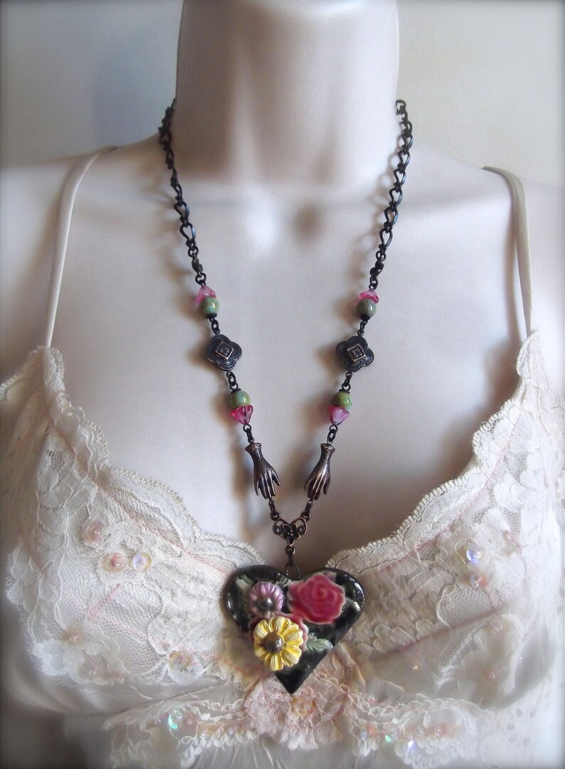 Pode incluir: Um colar de corrente preta com contas verdes e rosa e um pingente em forma de cora&ccedil;&atilde;o. O pingente &eacute; decorado com um desenho floral em tons de rosa, amarelo e verde.