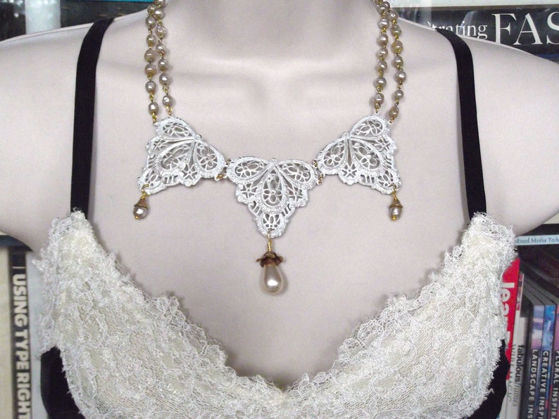 May include: A white lace necklace with a three-piece design and a pearl drop pendant. The necklace is made of delicate lace and features a gold chain.