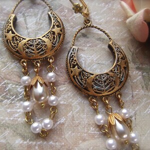 Puede incluir: Pendientes de tono dorado con un dise&ntilde;o de media luna y un motivo de &aacute;rbol. Los pendientes tienen una cadena colgante con cuentas de perlas blancas.