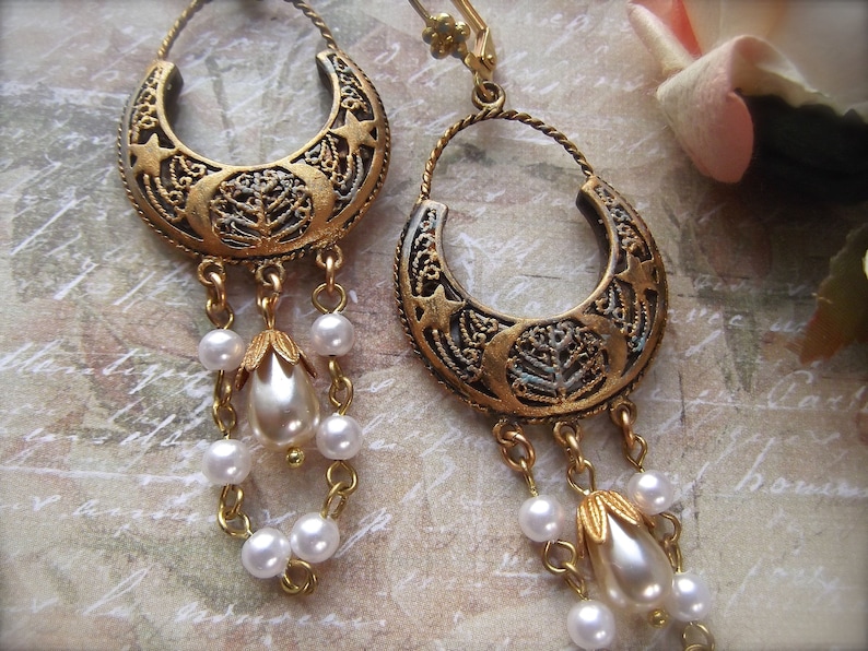 Puede incluir: Pendientes de tono dorado con un dise&ntilde;o de media luna y un motivo de &aacute;rbol. Los pendientes tienen una cadena colgante con cuentas de perlas blancas.