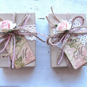 May include: Two small, brown gift boxes tied with twine and lace. Each box has a pink flower and a tag with the letter "M" on it.