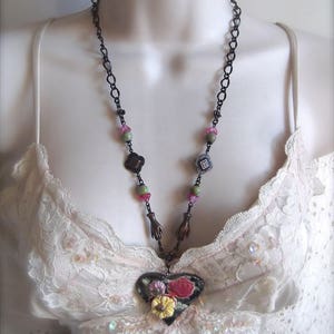 Pode incluir: Um colar de corrente preta com um pingente em forma de cora&ccedil;&atilde;o que apresenta um design floral em tons de rosa, verde e branco. O pingente &eacute; cercado por pequenos encantos em forma de m&atilde;os e quadrados.