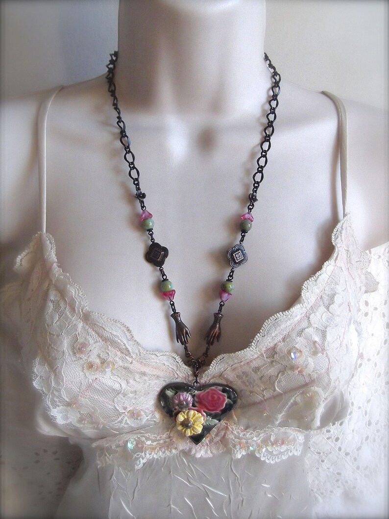 Pode incluir: Um colar de corrente preta com um pingente em forma de cora&ccedil;&atilde;o que apresenta um design floral em tons de rosa, verde e branco. O pingente &eacute; cercado por pequenos encantos em forma de m&atilde;os e quadrados.