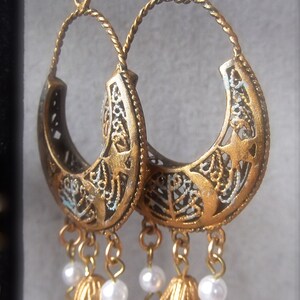 Puede incluir: Pendientes colgantes de tono dorado con un dise&ntilde;o de filigrana y detalles de perlas blancas.