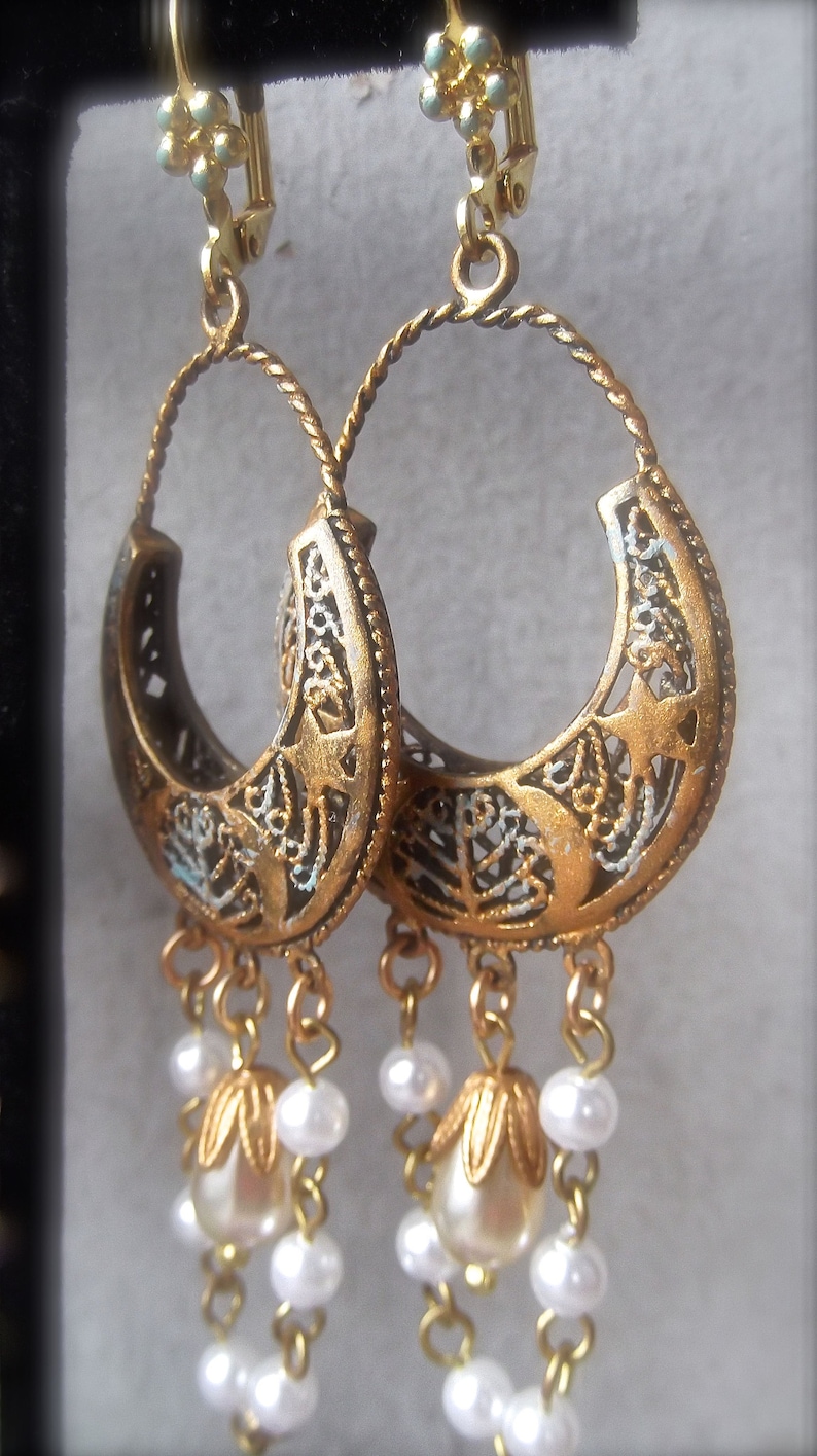 Puede incluir: Pendientes colgantes de tono dorado con un dise&ntilde;o de filigrana y detalles de perlas blancas.