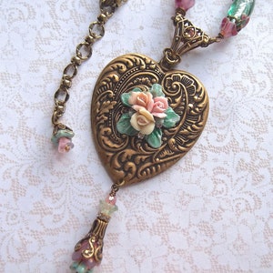 Puede incluir: Collar de estilo vintage con colgante en forma de coraz&oacute;n. El colgante, de color bronce, presenta un dise&ntilde;o floral en relieve y un racimo de rosas rosas y blancas. El collar incluye una cadena con cuentas decorativas en tonos rosa, verde y bronce.