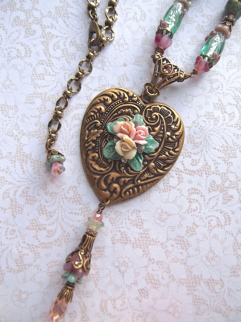 Puede incluir: Collar de estilo vintage con colgante en forma de coraz&oacute;n. El colgante, de color bronce, presenta un dise&ntilde;o floral en relieve y un racimo de rosas rosas y blancas. El collar incluye una cadena con cuentas decorativas en tonos rosa, verde y bronce.
