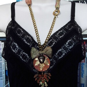 May include: A gold chain necklace with a large acorn pendant. The pendant is made of metal and has a brown and gold finish. The acorn is surrounded by leaves and a bow. The necklace is worn on a black lace top.