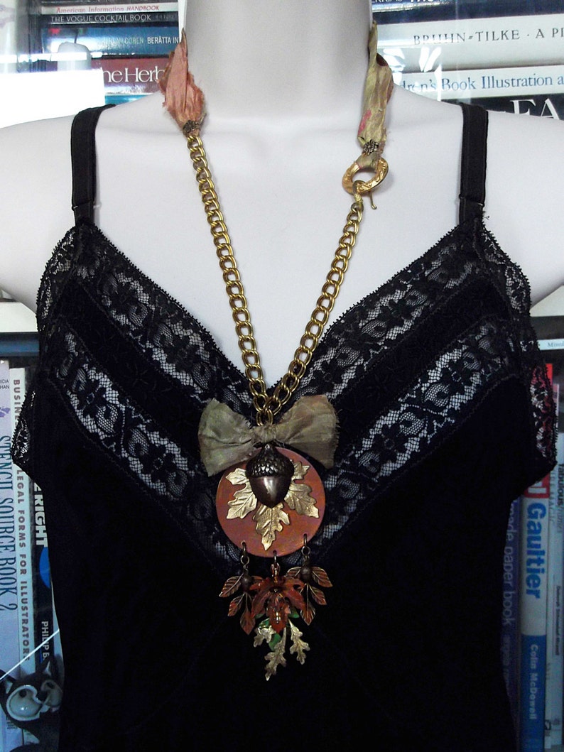 May include: A gold chain necklace with a large acorn pendant. The pendant is made of metal and has a brown and gold finish. The acorn is surrounded by leaves and a bow. The necklace is worn on a black lace top.
