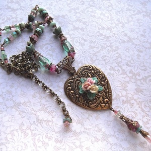 Puede incluir: Collar de estilo vintage con un colgante en forma de coraz&oacute;n adornado con flores. La cadena es de bronce y est&aacute; adornada con cuentas rosas, verdes y transparentes. El colgante es de bronce y tiene un dise&ntilde;o floral.