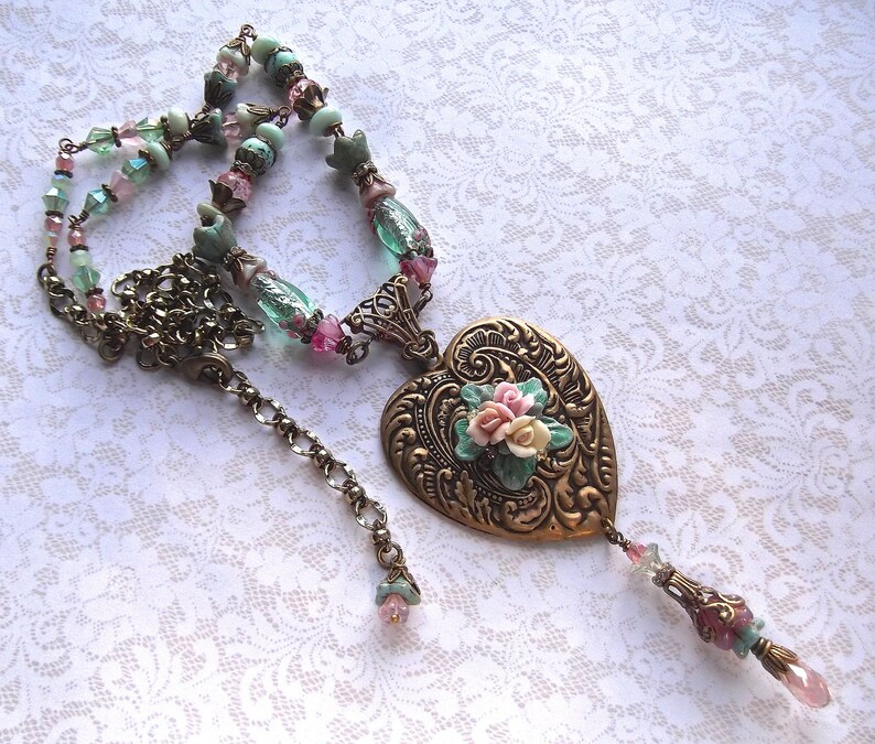 Puede incluir: Collar de estilo vintage con un colgante en forma de coraz&oacute;n adornado con flores. La cadena es de bronce y est&aacute; adornada con cuentas rosas, verdes y transparentes. El colgante es de bronce y tiene un dise&ntilde;o floral.