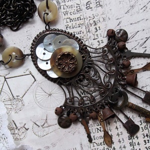 May include: A vintage-inspired necklace with a large, ornate pendant featuring a cluster of mother-of-pearl buttons and a brass button with a floral design. The pendant is attached to a chain with a variety of charms, including keys, bells, and spoons.