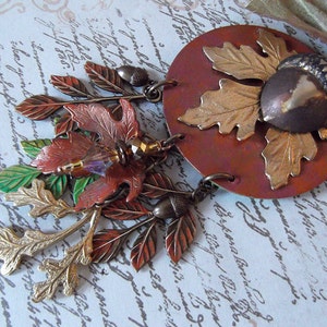 May include: A copper and gold necklace with a large acorn pendant and multiple leaf charms. The leaves are in shades of green, gold, and brown. The necklace is on a white background with black writing.