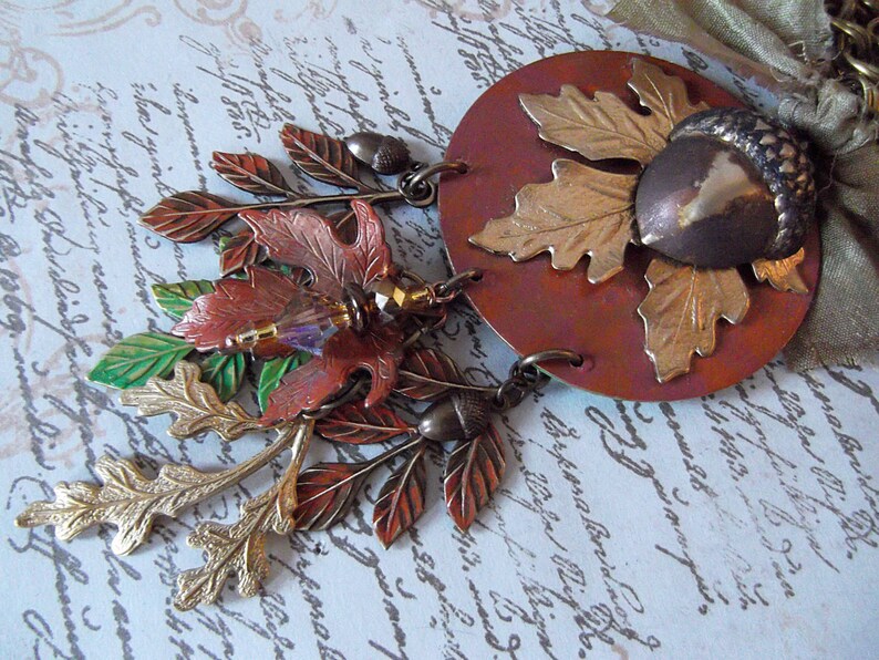 May include: A copper and gold necklace with a large acorn pendant and multiple leaf charms. The leaves are in shades of green, gold, and brown. The necklace is on a white background with black writing.