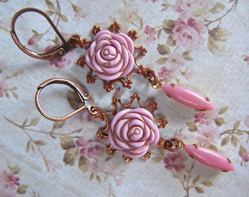 May include: A pair of pink rose earrings with copper-toned metal accents. The earrings feature a delicate floral design and are perfect for adding a touch of vintage charm to any outfit.