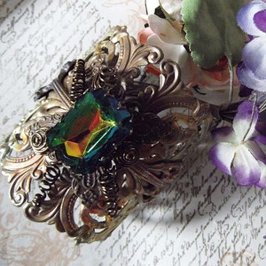 May include: A gold-toned metal hair comb with an ornate design featuring a large iridescent green gemstone. The comb is set against a background of white paper with handwritten text.