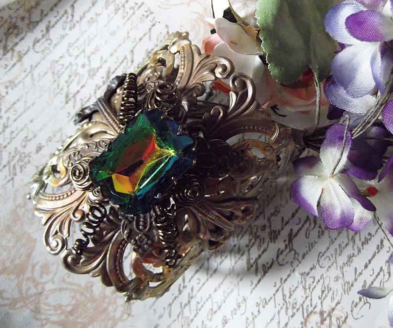 May include: A gold-toned metal hair comb with an ornate design featuring a large iridescent green gemstone. The comb is set against a background of white paper with handwritten text.