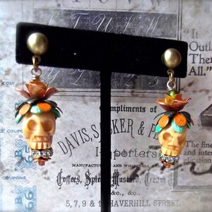 May include: A pair of skull-shaped earrings with orange, turquoise, and white enamel details. The earrings are hanging from a black display stand. The text "Outlasts Them All." is visible in the background.